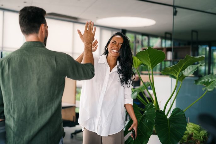 Two colleagues sharing a high-five in a bright, modern office space surrounded by greenery.