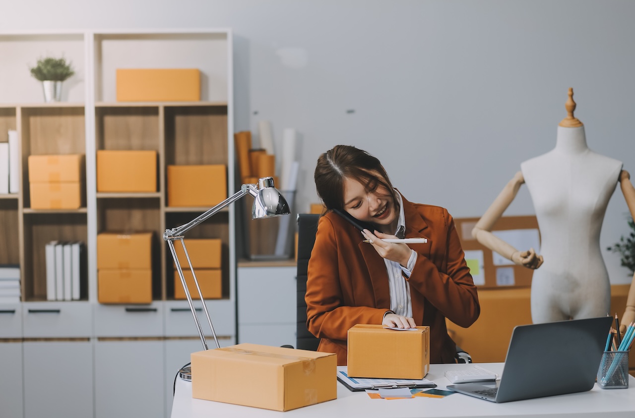 Small business owner multitasking at desk while preparing packages and speaking with a supplier on the phone.