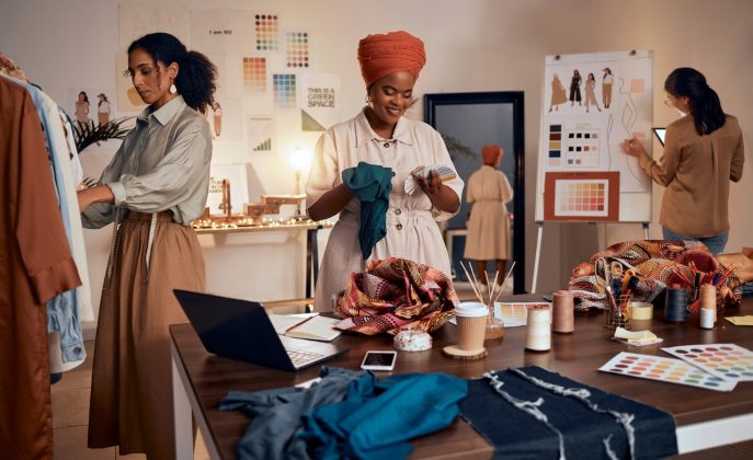 Three women collaborating in a creative fashion design studio, surrounded by fabrics, colour palettes, and sketches