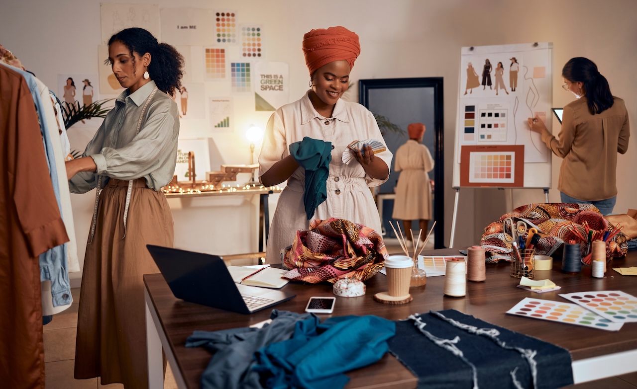 Three women collaborating in a creative fashion design studio, surrounded by fabrics, colour palettes, and sketches
