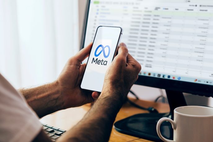Person holding a smartphone displaying the Meta logo in front of a desktop screen with analytics data.