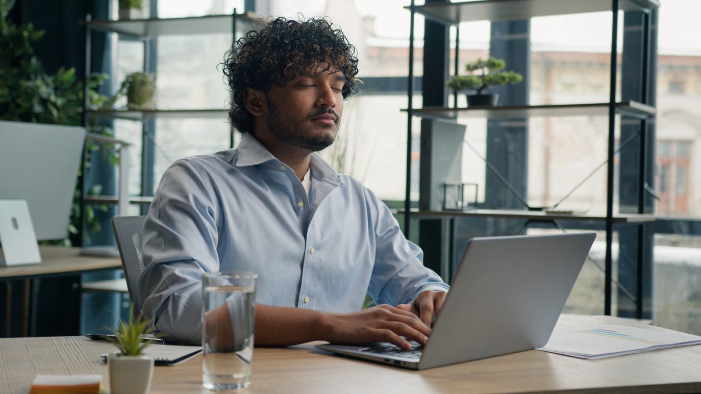 Calm business owner sitting with eyes closed at laptop, practicing mindfulness in the office.