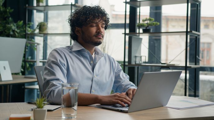 Calm business owner sitting with eyes closed at laptop, practicing mindfulness in the office.
