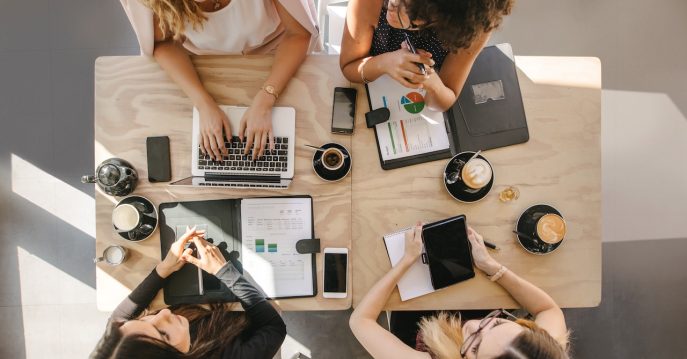 Top-down view of diverse women working on marketing strategies at a coffee-fueled business meeting.