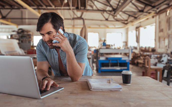 Man on phone and laptop in workshop reviewing paperwork