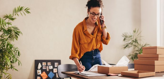 Smiling female entrepreneur on the phone while preparing product packages and taking notes at a desk.