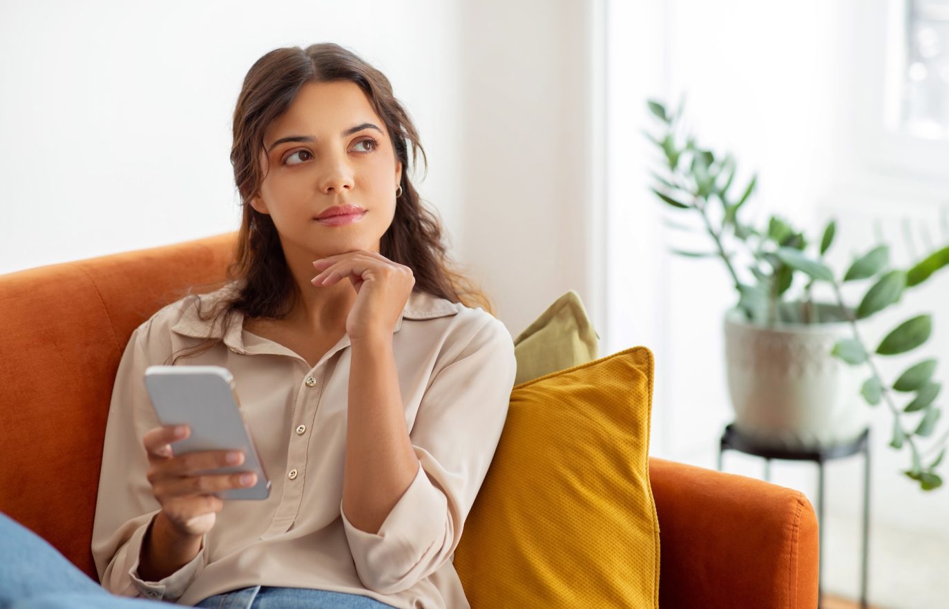 Thoughtful young woman holding smartphone while sitting on couch at home
