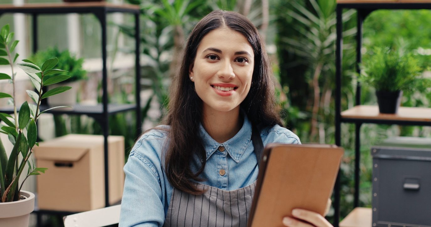 Smiling female entrepreneur in apron holding digital tablet in plant-filled workspace