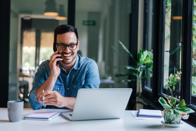 Smiling businessman on phone call while working on laptop at bright office desk