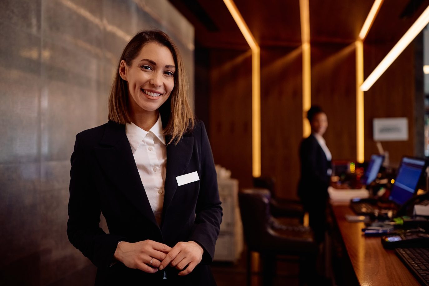 Hotel receptionist in uniform smiling at the front desk of a modern hotel lobby.