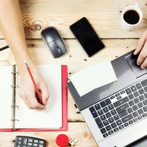 Overhead view of hands working with a laptop, notebook, phone, and coffee on a wooden desk