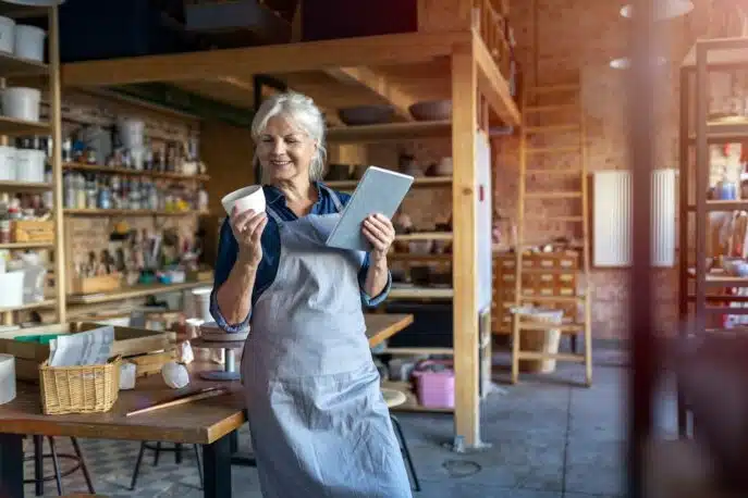 Smiling woman in an apron holding a tablet and ceramic cup in a creative studio