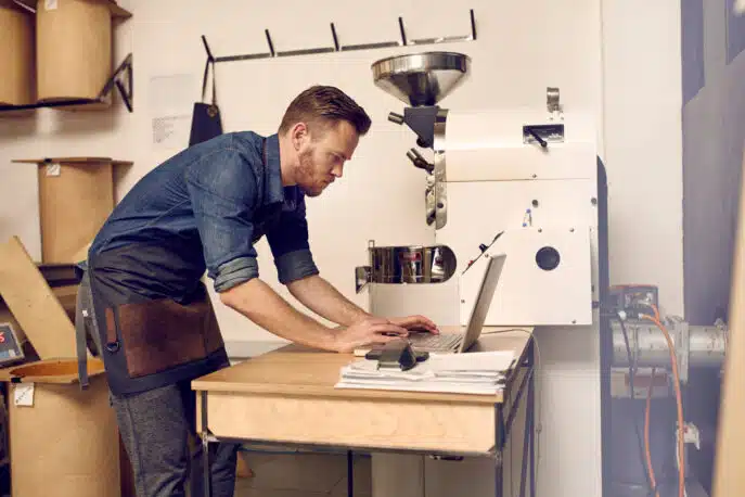 Small business owner using laptop beside industrial coffee roasting machine