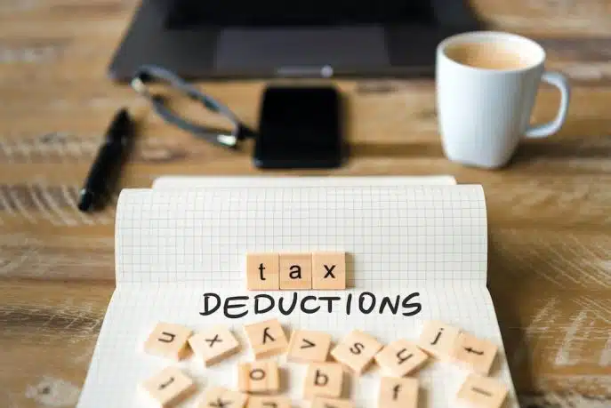 Notebook page with the words “tax deductions” spelled out using wooden letter tiles on a desk with a coffee cup, glasses, and a laptop