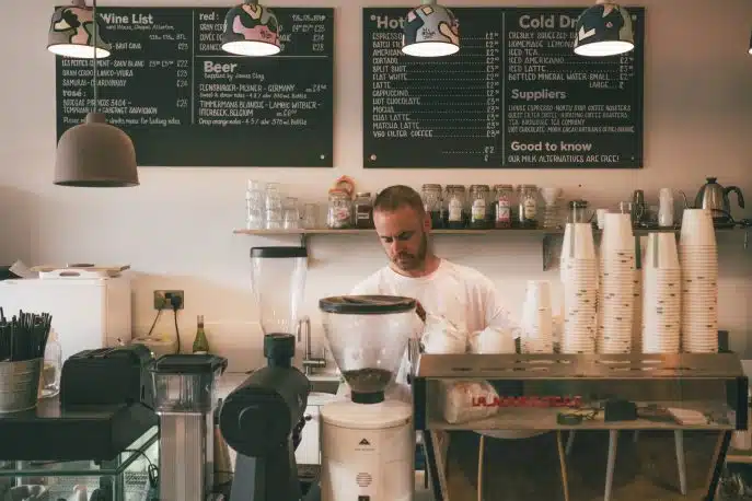 Barista working behind the counter in a modern café with coffee machines, takeaway cups, and a chalkboard menu