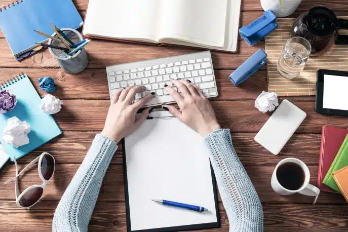 Person typing on a wireless keyboard at a cluttered desk with notebooks, pens, and coffee