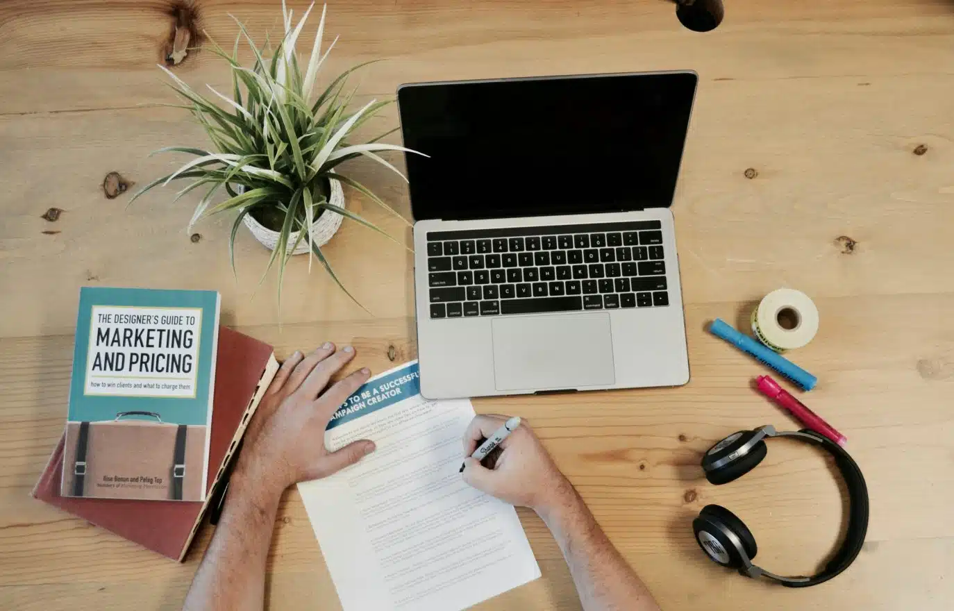 Overhead view of a person working at a wooden desk with a laptop, marketing book, and campaign notes.