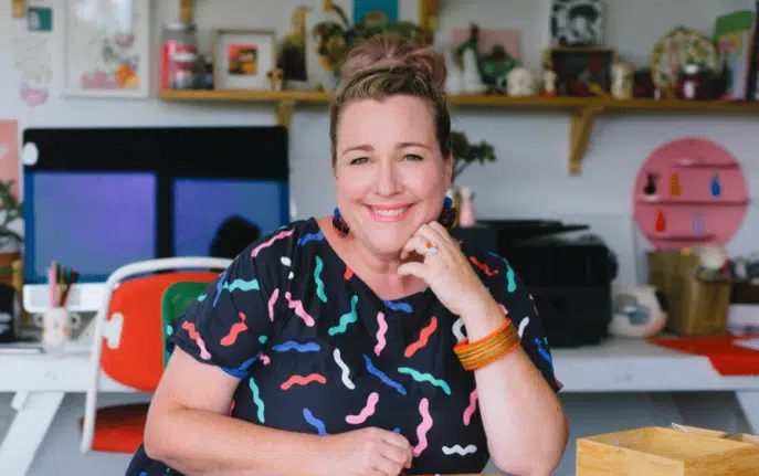 Smiling woman sitting in a colorful home office with creative decor and artwork behind her