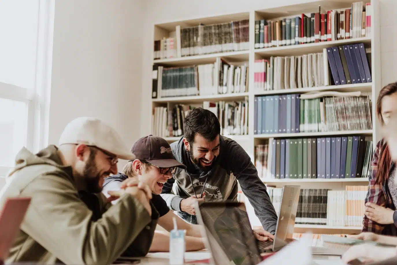 Group of young adults collaborating and laughing while working on laptops in a library or shared workspace.