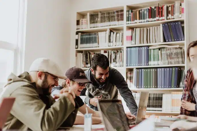 Group of young adults collaborating and laughing while working on laptops in a library or shared workspace.