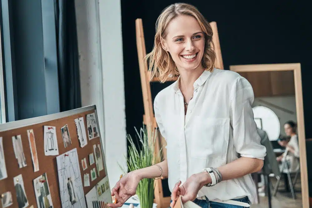 Smiling woman standing in a modern studio beside a corkboard with fashion sketches and fabric swatches