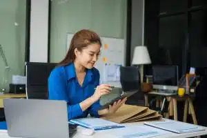 Businesswoman in blue shirt reviewing documents on a tablet at a desk with files and a laptop.