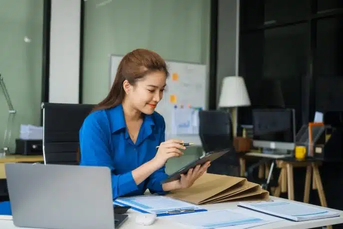 Businesswoman in blue shirt reviewing documents on a tablet at a desk with files and a laptop.