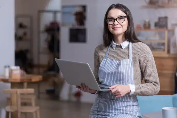 Smiling business owner in apron holding a laptop inside her shop