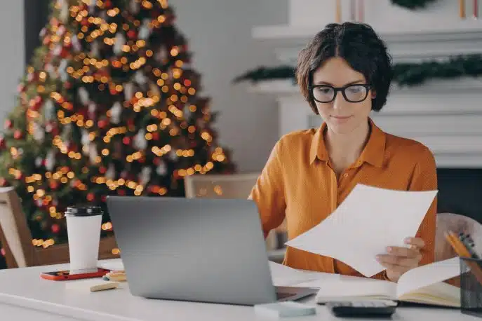 Woman working on documents at a laptop in a festive home office with a decorated Christmas tree in the background.