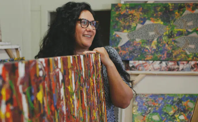 Smiling artist holding a large colorful canvas with abstract and nature-themed paintings in the background