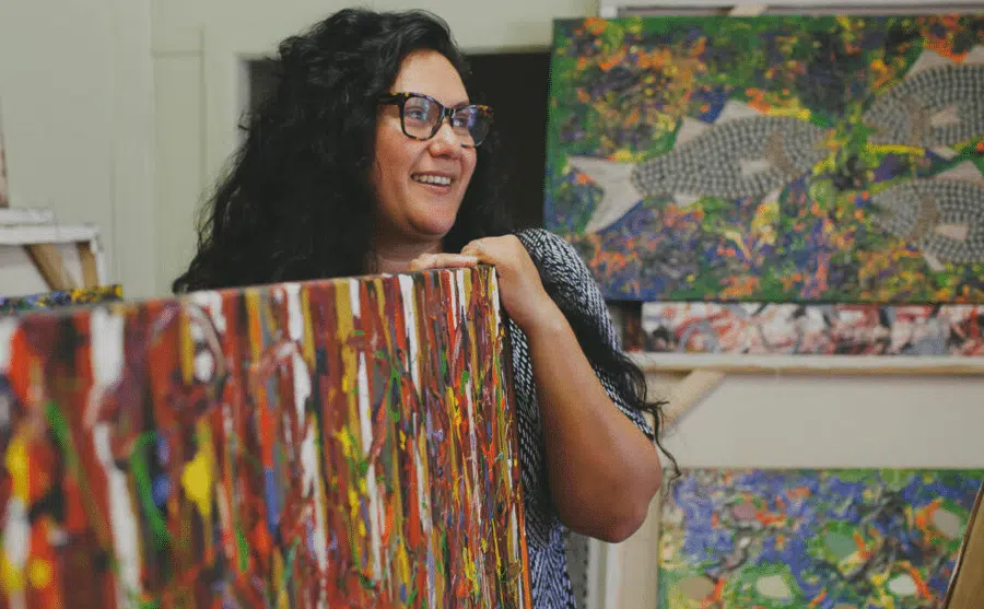 Smiling artist holding a large colorful canvas with abstract and nature-themed paintings in the background