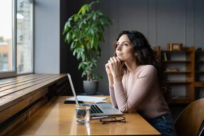 Thoughtful businesswoman sitting at desk with laptop, looking out the window.