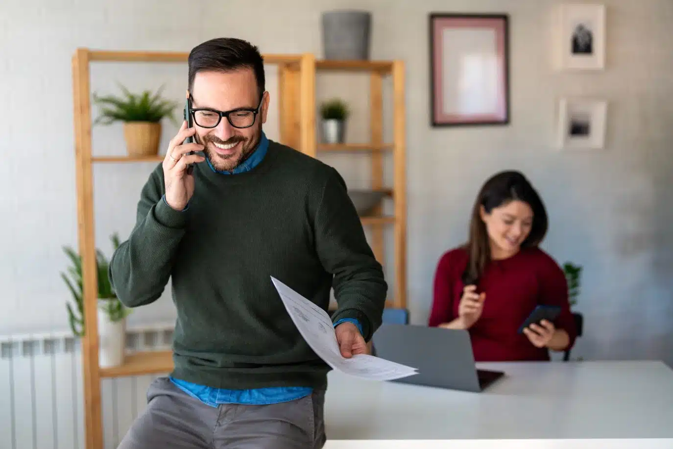 Smiling man on phone holding documents with woman using a tablet in the background.