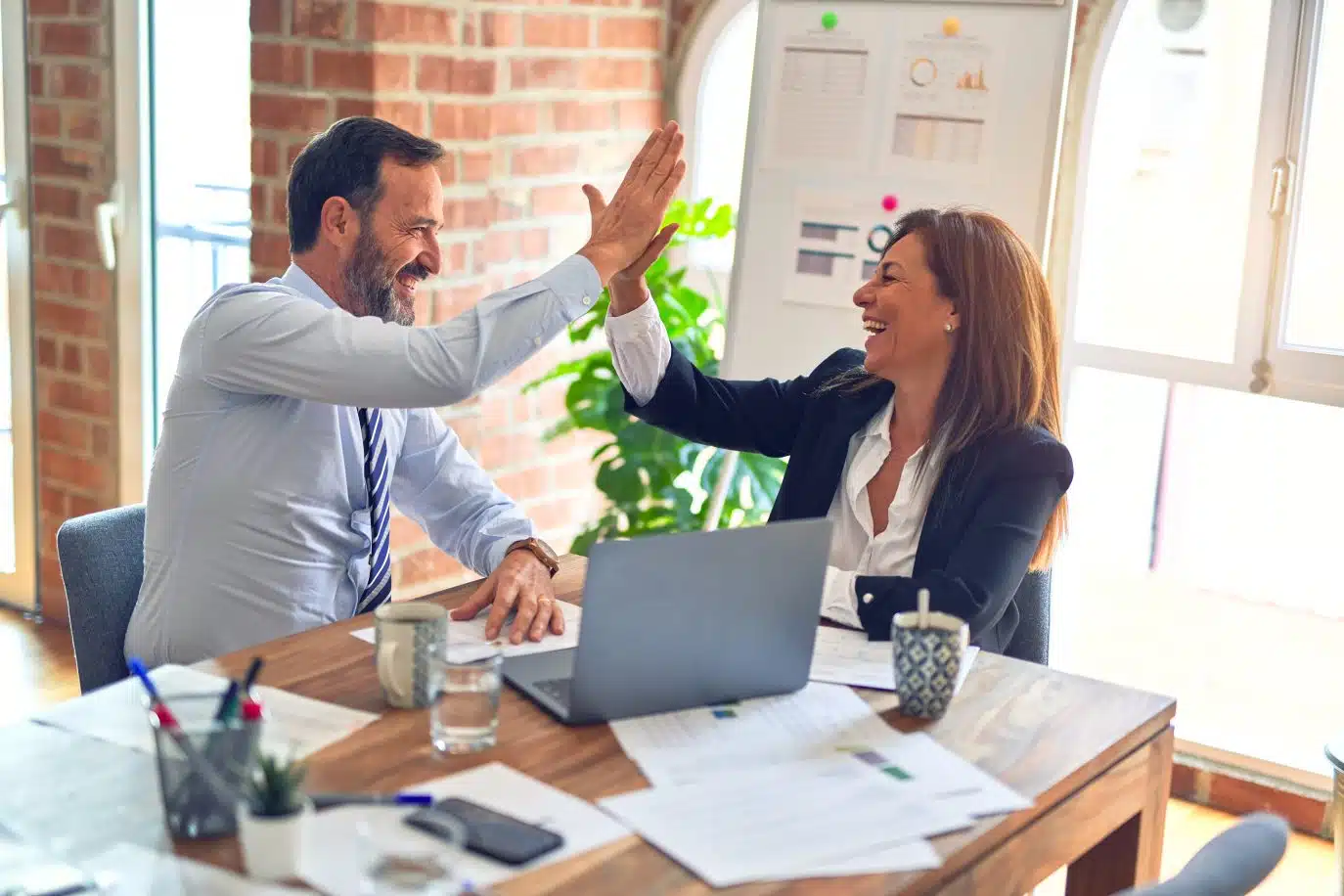 Two business professionals sitting at a desk give each other a high five in celebration, smiling in a brightly lit office space.