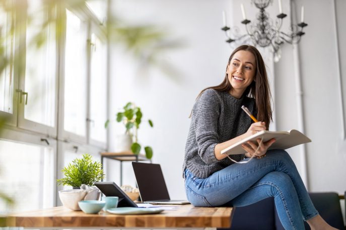 Smiling woman writing notes while sitting on a table with a laptop