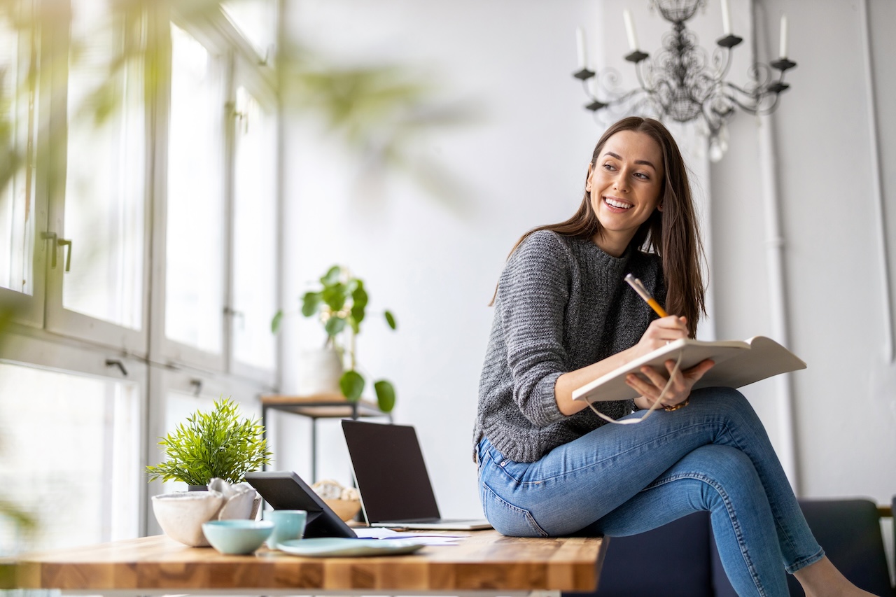 Smiling woman writing notes while sitting on a table with a laptop